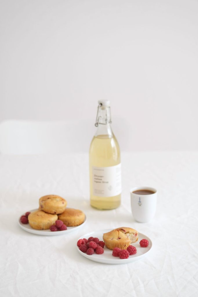 Frische Himbeeren und Himbeer-Muffins mit einer Flasche Zitronensirup und einem Becher Kaffee auf einem weiß gedeckten Tisch.